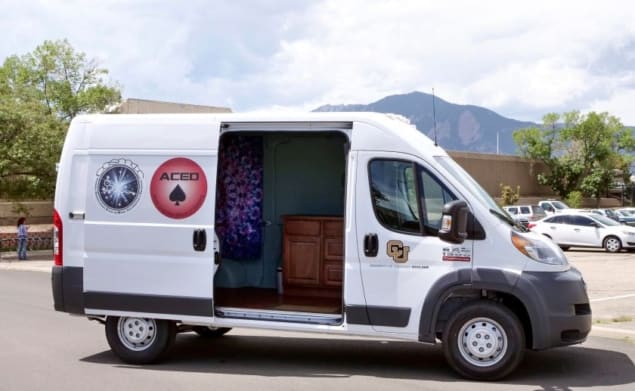 Photo of a white van used as a mobile testing centre for measuring THC in the breath of cannabis users. The van door is open, revealing a psychedelic purple wall hanging and a chest of drawers inside