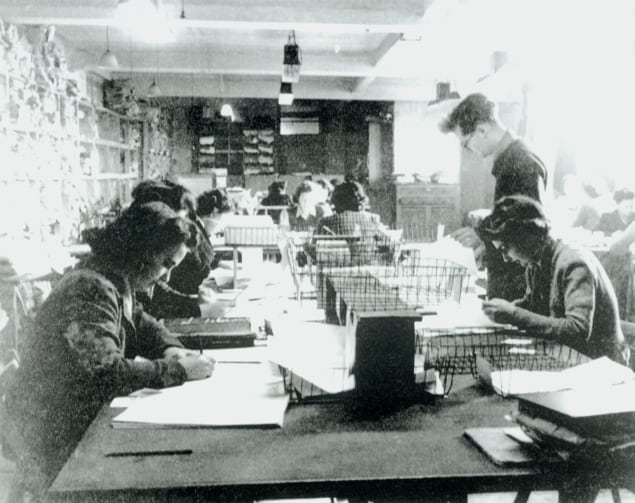 Black and white photo of men and women bent over paperwork at a long wooden table