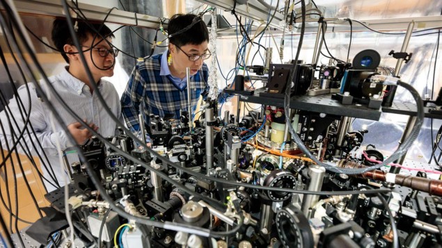 Photo of Zhendong Zhang and Cheng Chin in their lab at Chicago. They're looking through a tangle of wires at an optical bench full of components with a vacuum chamber at its heart