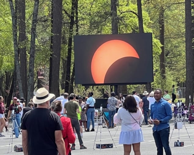 Uma foto de uma multidão de pessoas em frente a uma tela de projeção gigante representando a sombra da Lua passando na frente do Sol